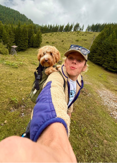 Hiker making a selfie with a small dog in a backpack on a mountain trail.